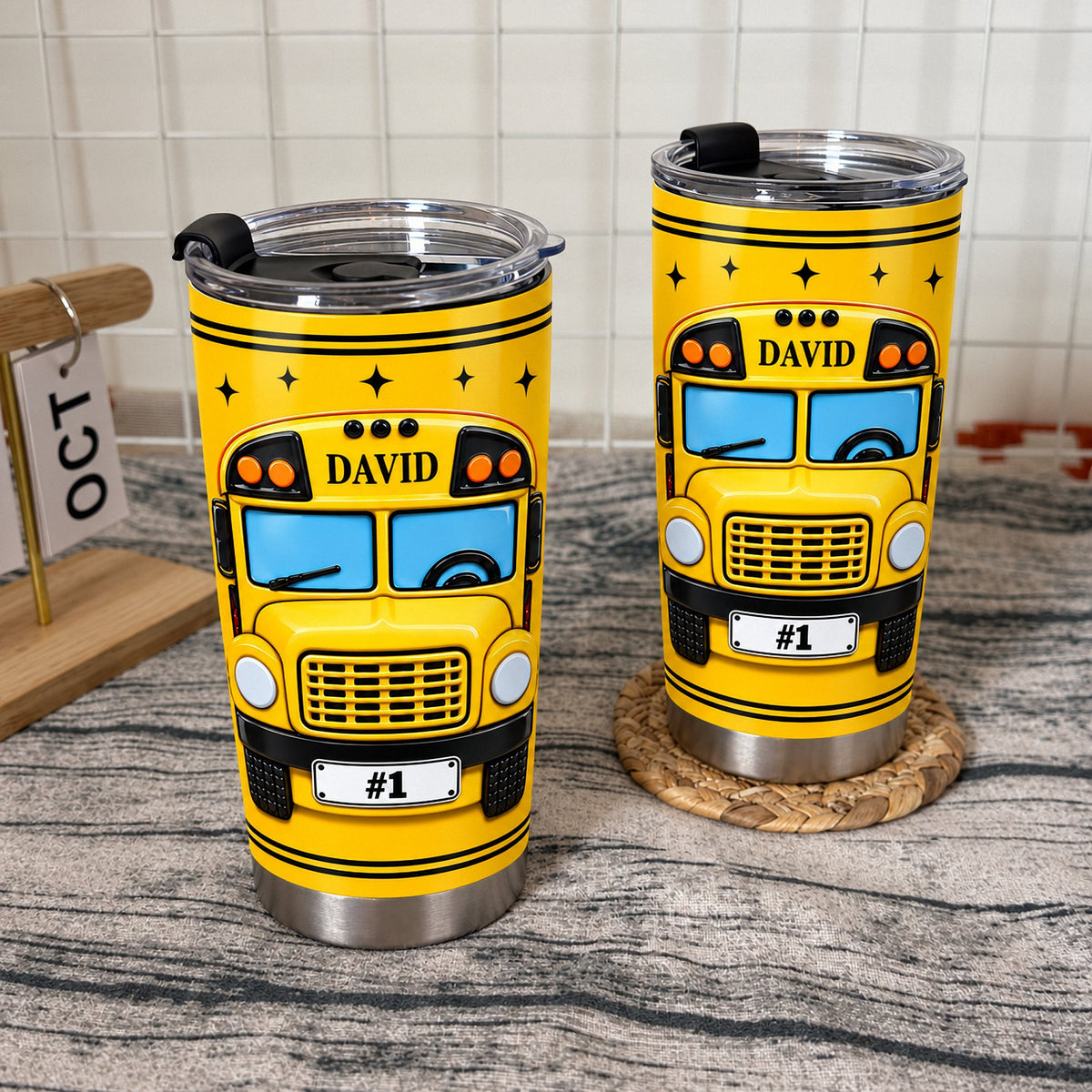 Two yellow school bus driver tumblers in stainless steel with custom name displayed side-by-side on a rustic wooden surface.