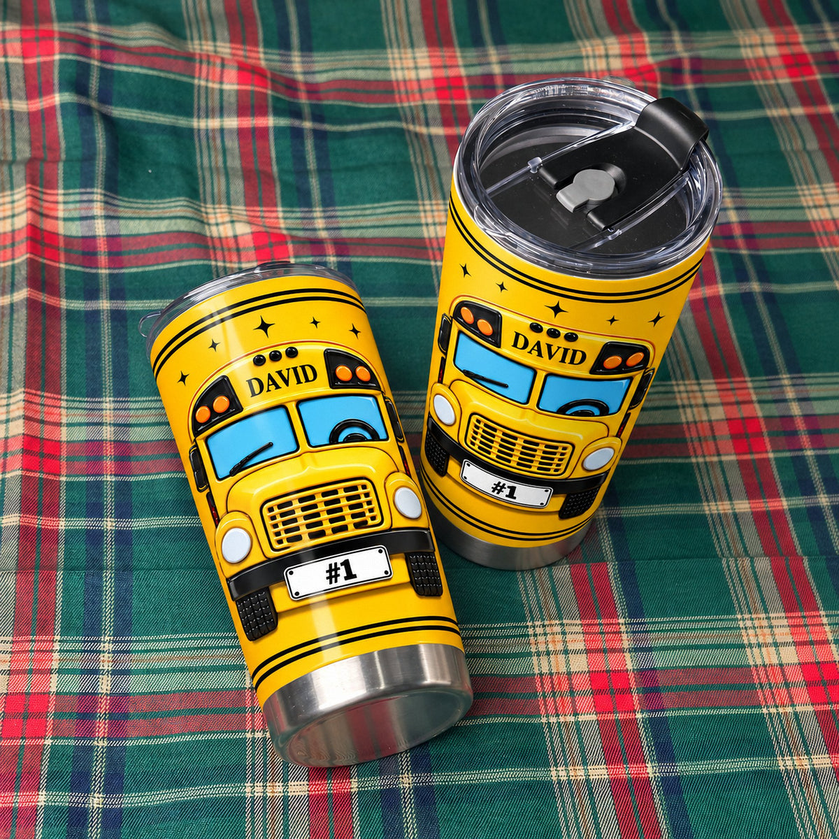 Two personalized school bus driver tumblers resting on a green and red plaid fabric background with visible lids.