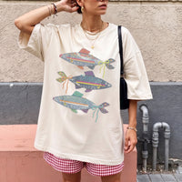 A woman wearing an off-white oversized t-shirt with three denim fish graphics and red gingham shorts.