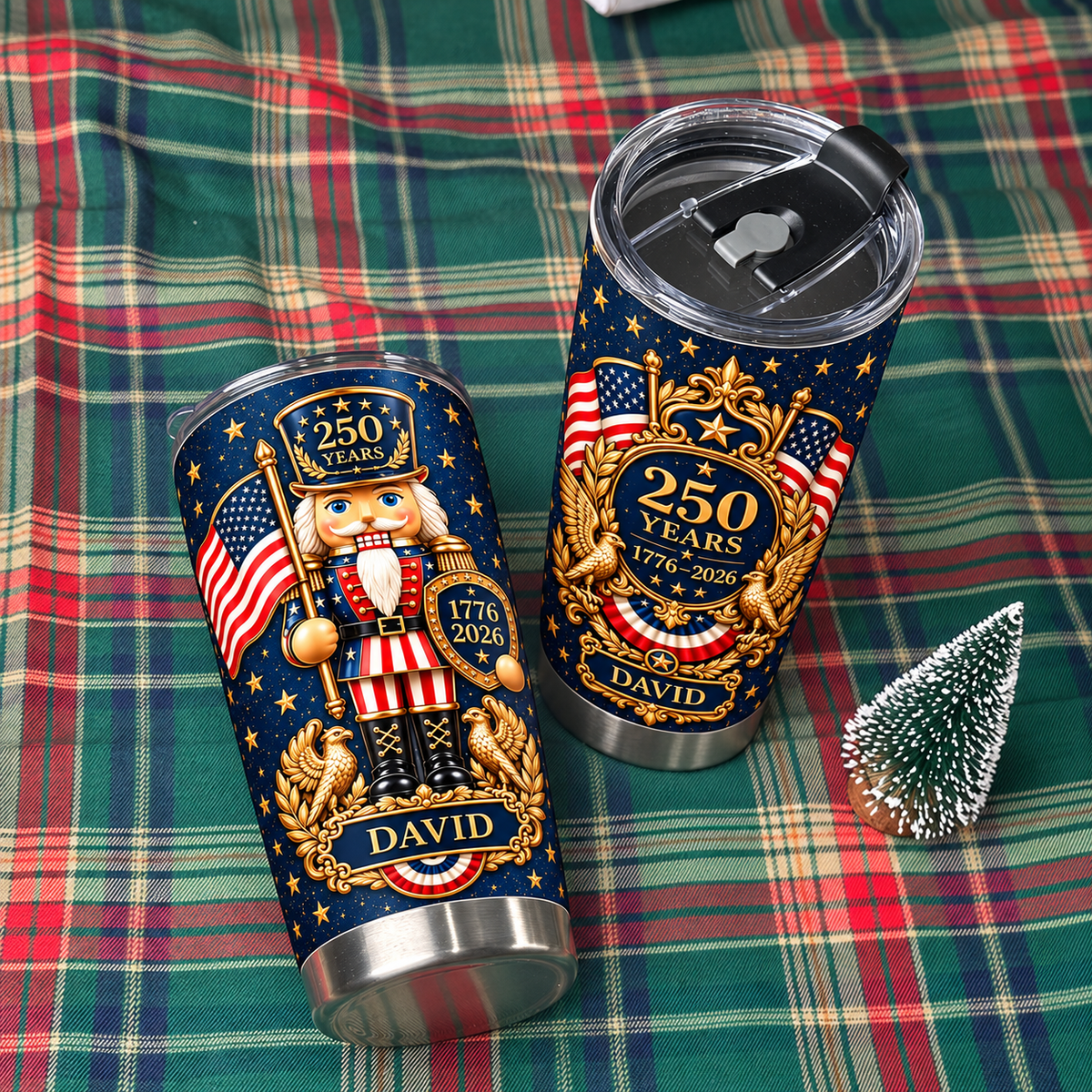 Two patriotic nutcracker tumblers resting on a green and red plaid blanket beside a tiny decorative pine tree.