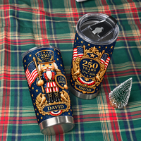 Two patriotic nutcracker tumblers resting on a green and red plaid blanket beside a tiny decorative pine tree.