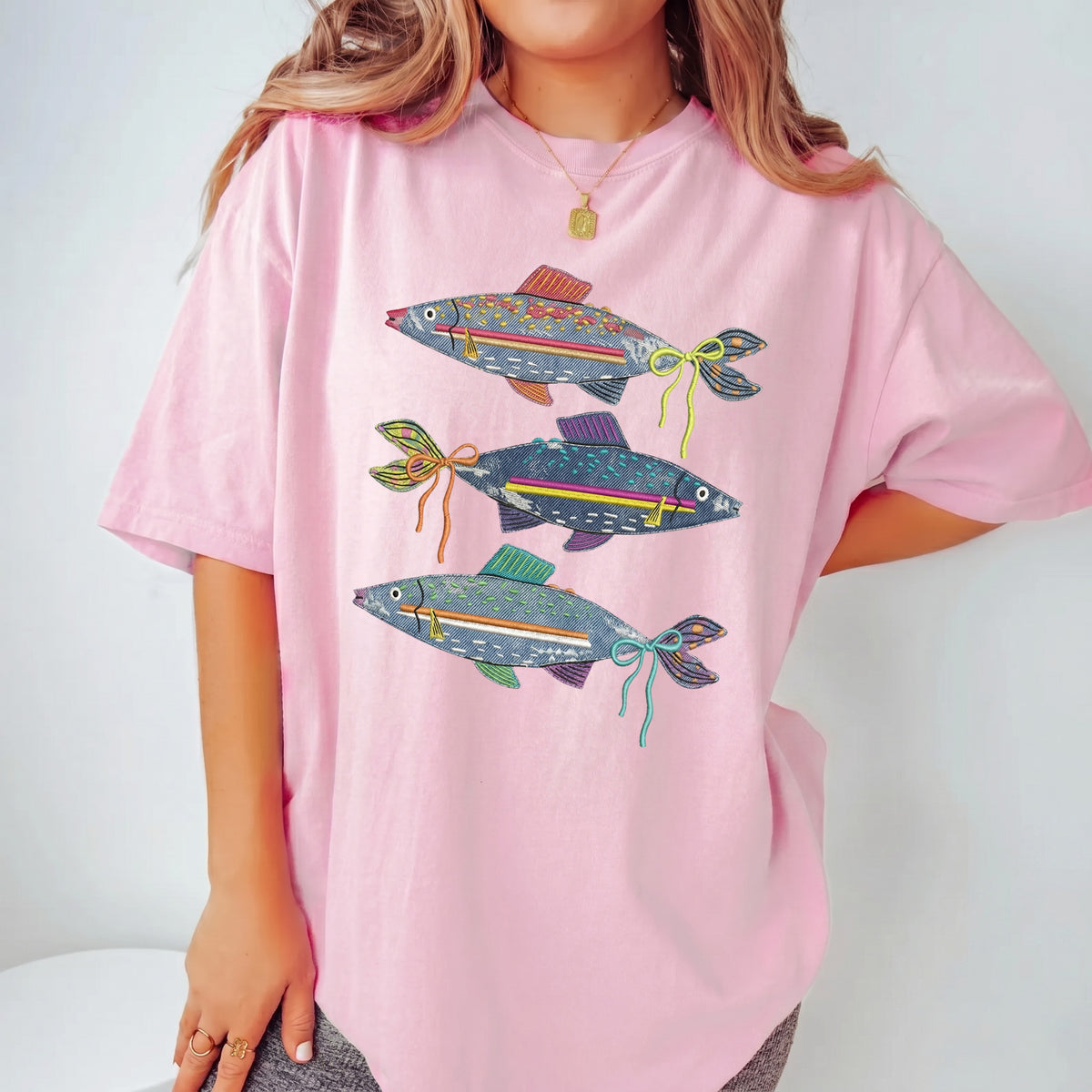 A light pink oversized t-shirt with a denim fish graphic, worn by a woman with layered gold jewelry.