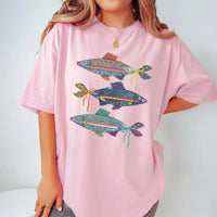 A light pink oversized t-shirt with a denim fish graphic, worn by a woman with layered gold jewelry.