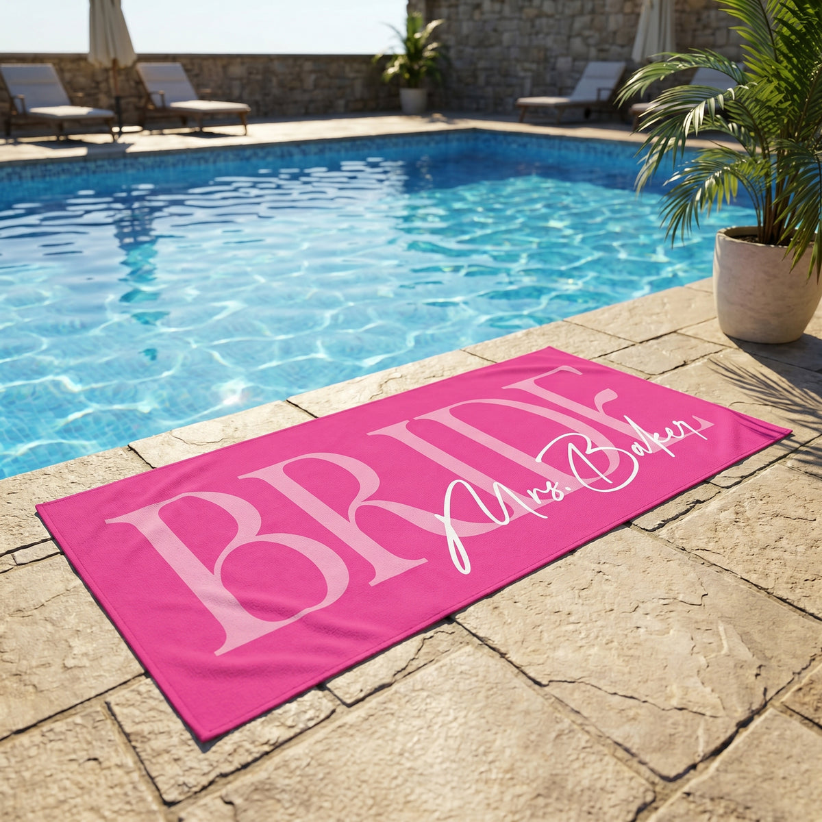 Hot pink beach towel with white BRIDE text and Mrs. Baker script laid on stone patio beside outdoor pool.