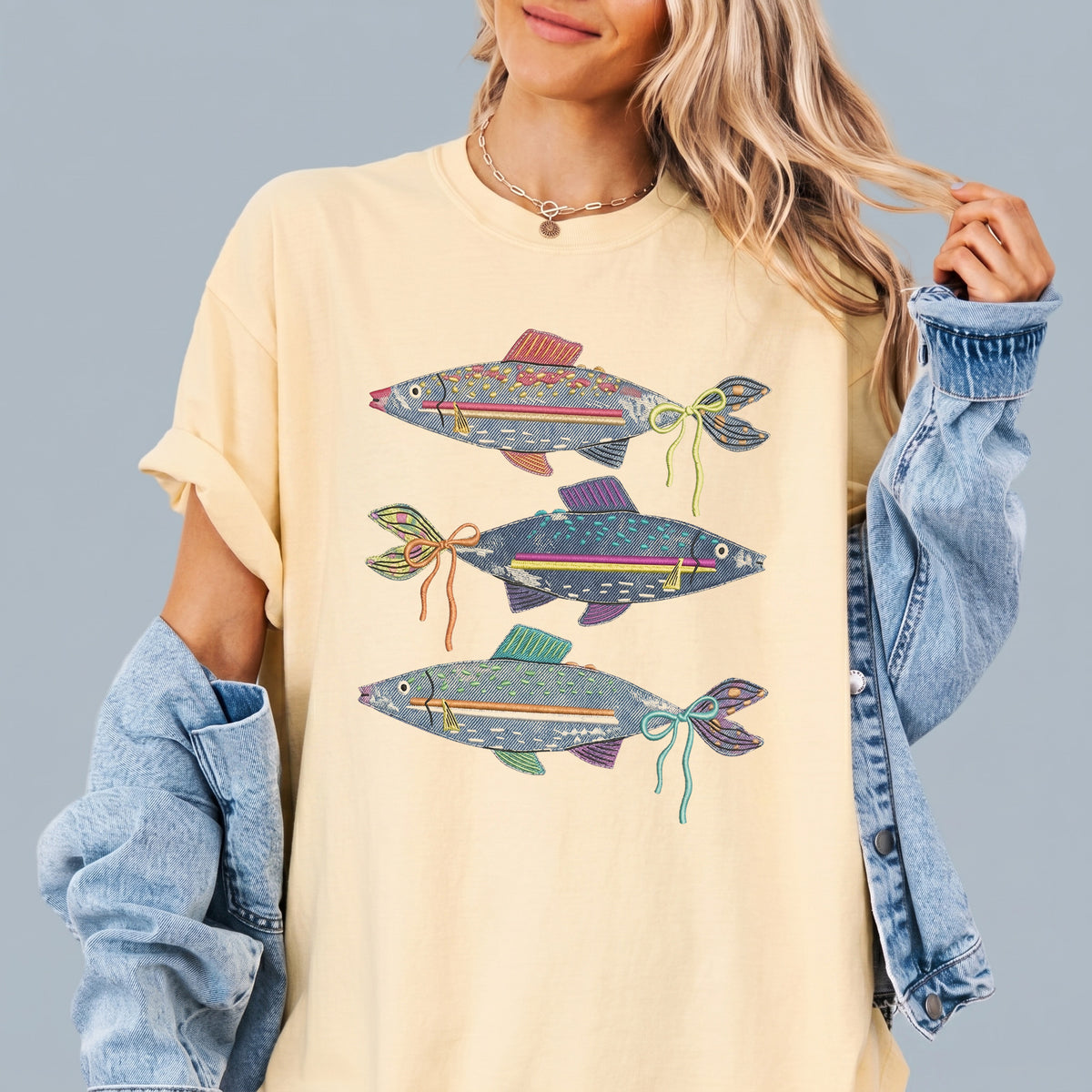 A person wearing a light yellow oversized graphic t-shirt featuring three denim fish with colorful bows, layered under a