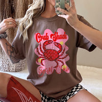 A woman posing in a brown graphic t-shirt with a crab design, wearing patterned shorts and red cowgirl boots.