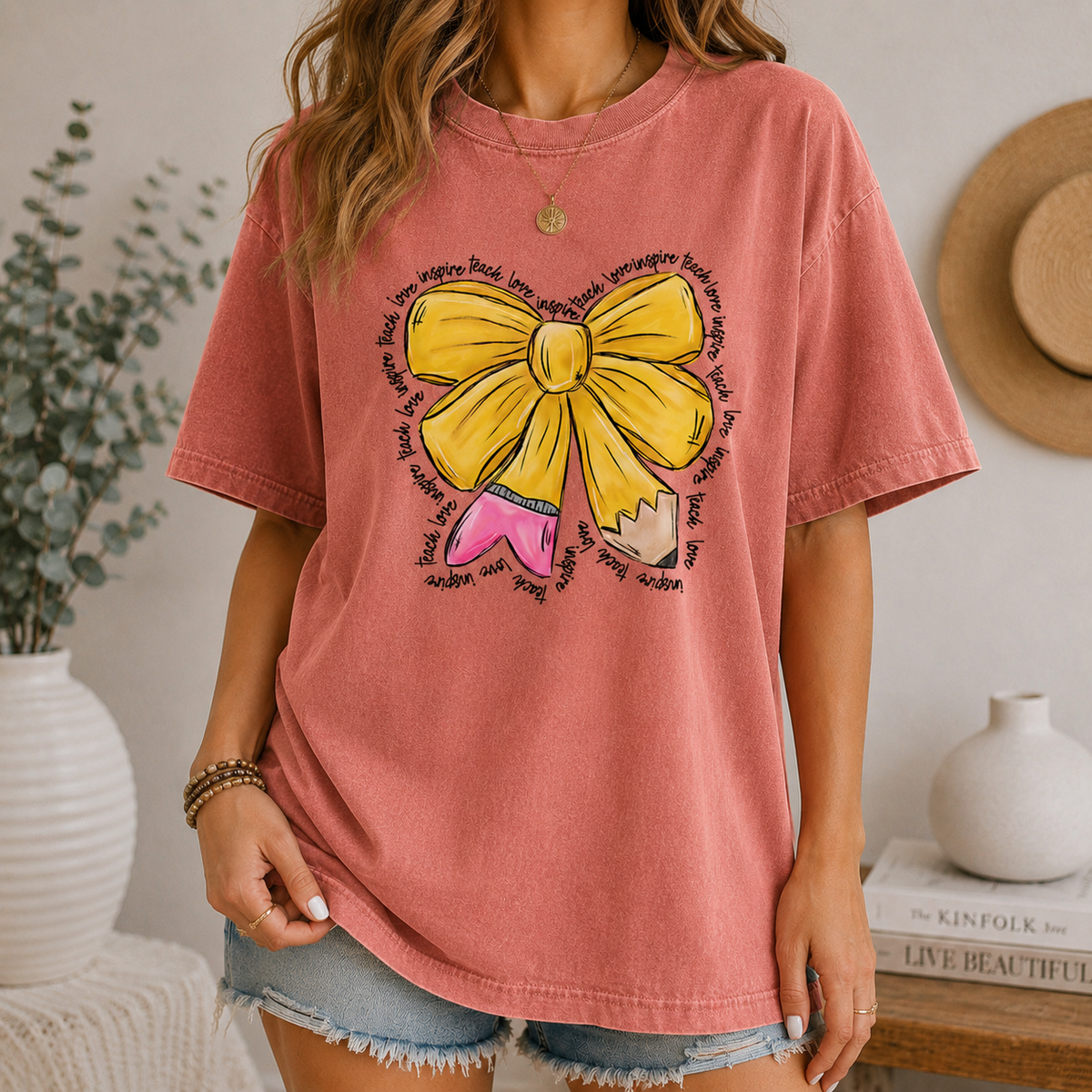 A woman wearing a terracotta t-shirt featuring a yellow bow and pencil graphic, paired with denim shorts.