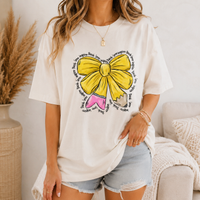 A woman wearing an off white t-shirt with a yellow bow and pencil graphic, paired with denim shorts.