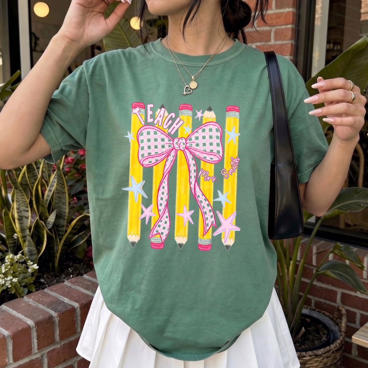 An olive green t-shirt with a pink bow and pencil graphic, paired with a white pleated skirt, worn outdoors.