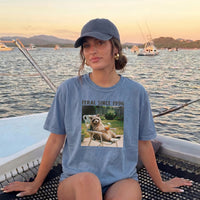 A woman wearing a blue-grey t-shirt with the raccoon meme graphic sits on a boat with water and distant mountains in the