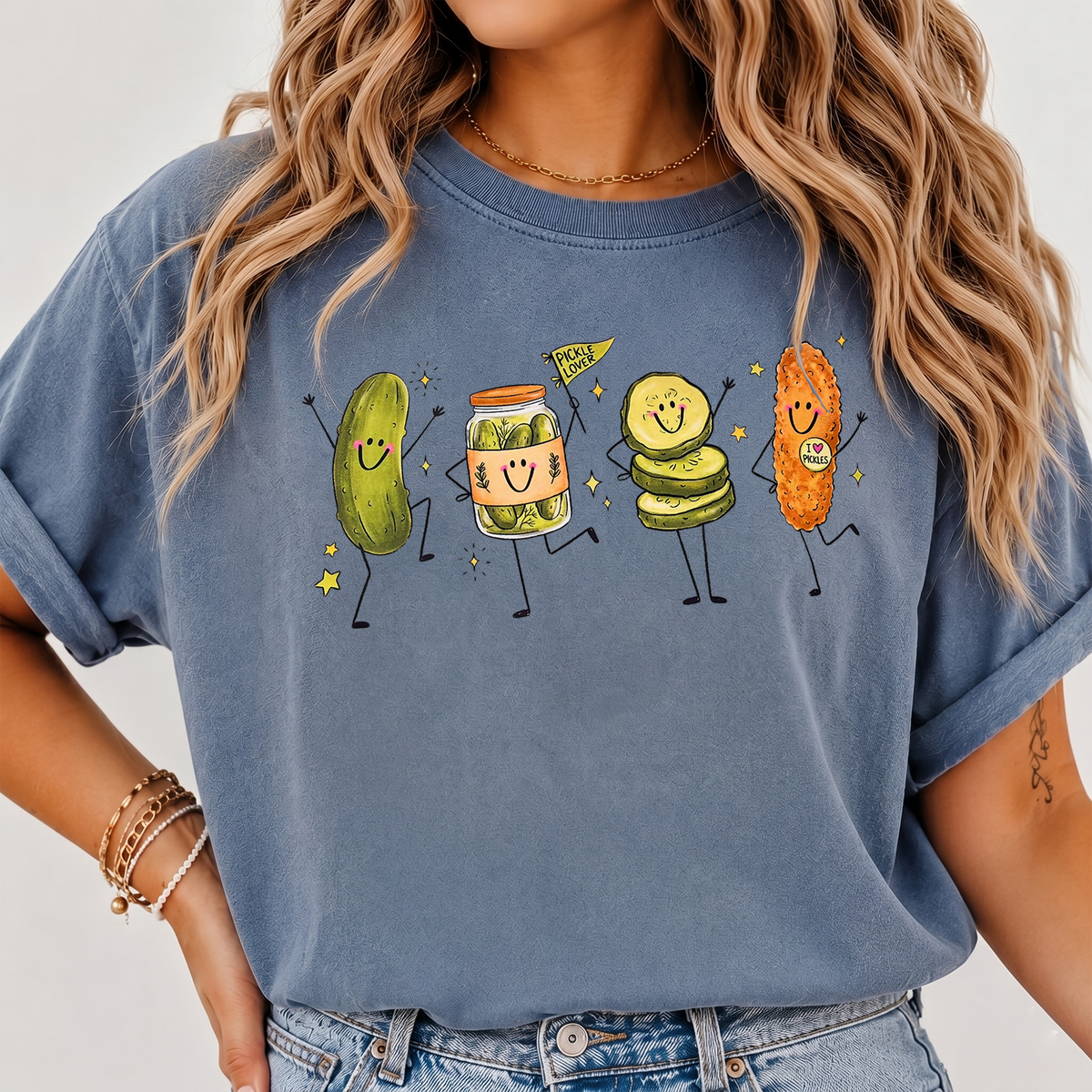 A woman wearing a blue-grey T-shirt featuring a cartoon dancing pickle graphic and casual jeans.