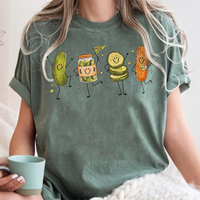 A woman sitting with a teal mug, wearing a dark green T-shirt with a playful dancing pickle graphic design.