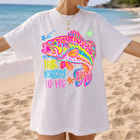A person on a sandy beach wearing a white t-shirt printed with a neon pink and blue bass fish.
