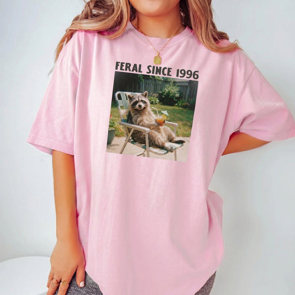 A woman models a casual pink t-shirt with a vintage raccoon meme graphic and the text Feral Since 1996 printed across the