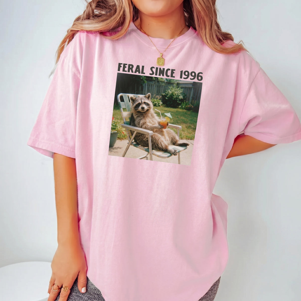 A woman models a casual pink t-shirt with a vintage raccoon meme graphic and the text Feral Since 1996 printed across the
