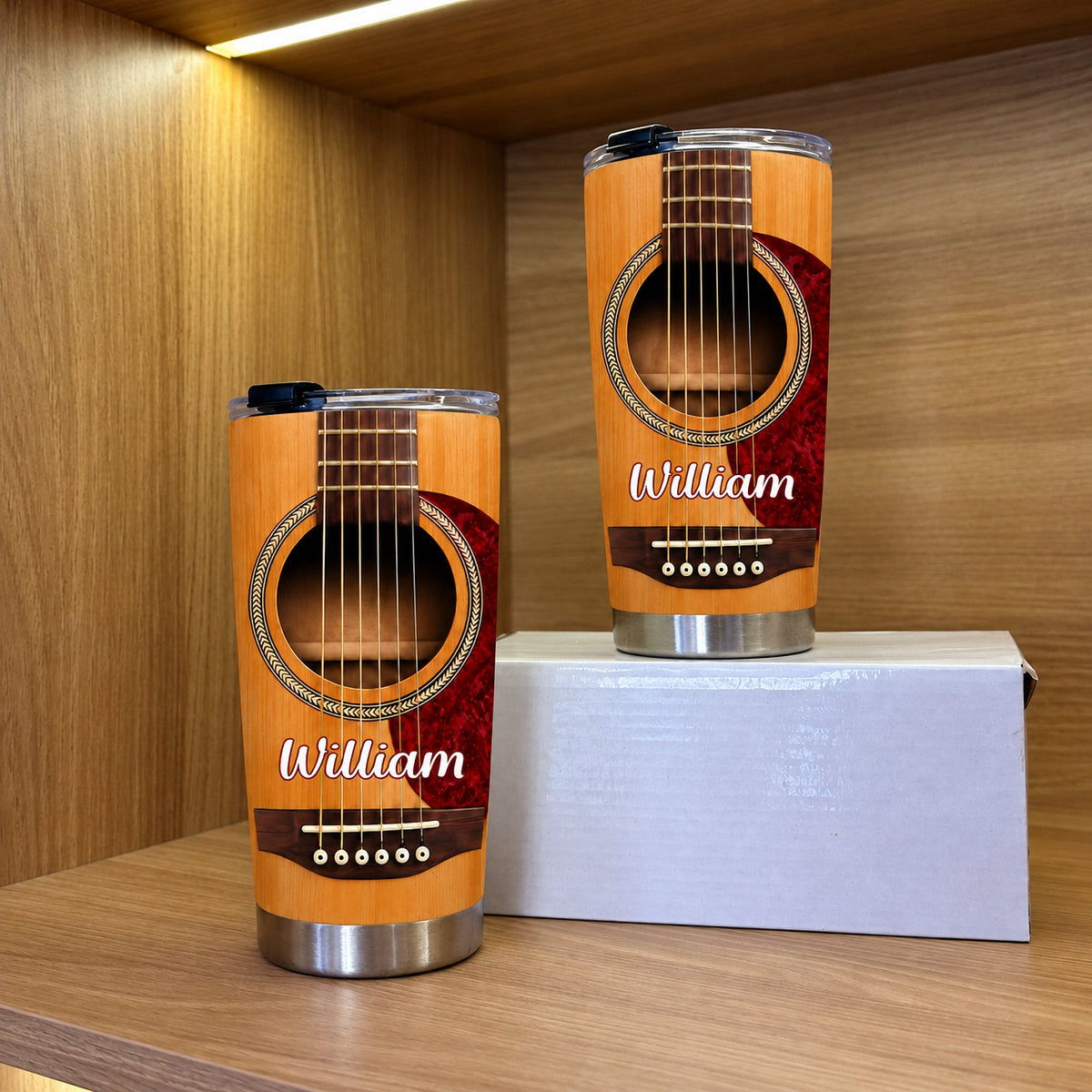 Two personalized acoustic guitar travel mugs displayed on a wooden shelf next to a gift box for guitarists.