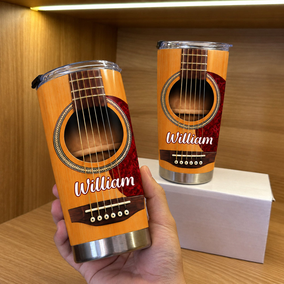 A person holding a personalized acoustic guitar tumbler next to another tumbler and white box on a wooden shelf.