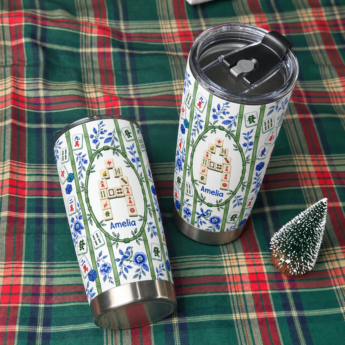 Two stainless steel travel tumblers featuring chinoiserie style designs rest on a plaid holiday blanket.