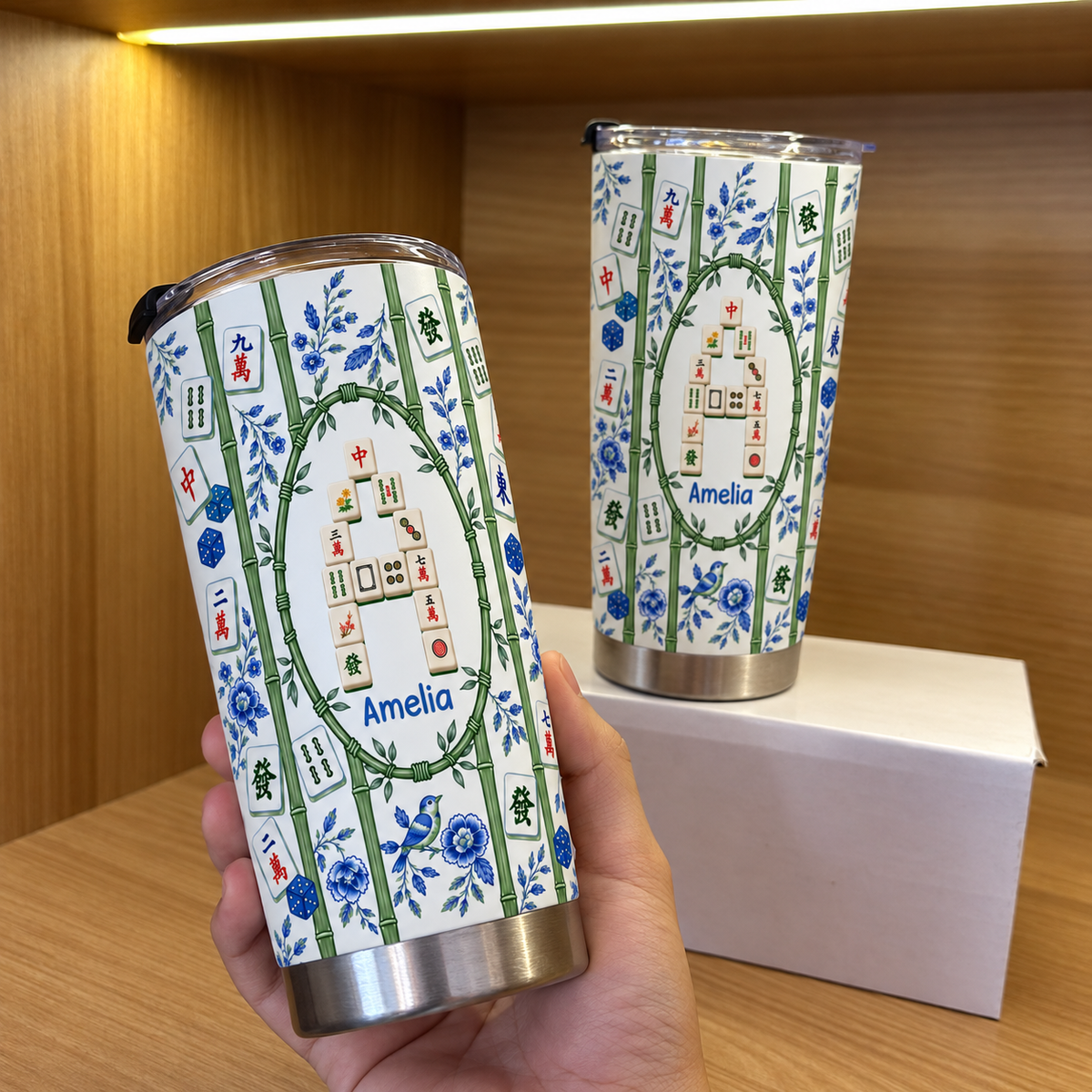 A hand holds a personalized mahjong tumbler with a taller travel cup sitting on a box behind it.