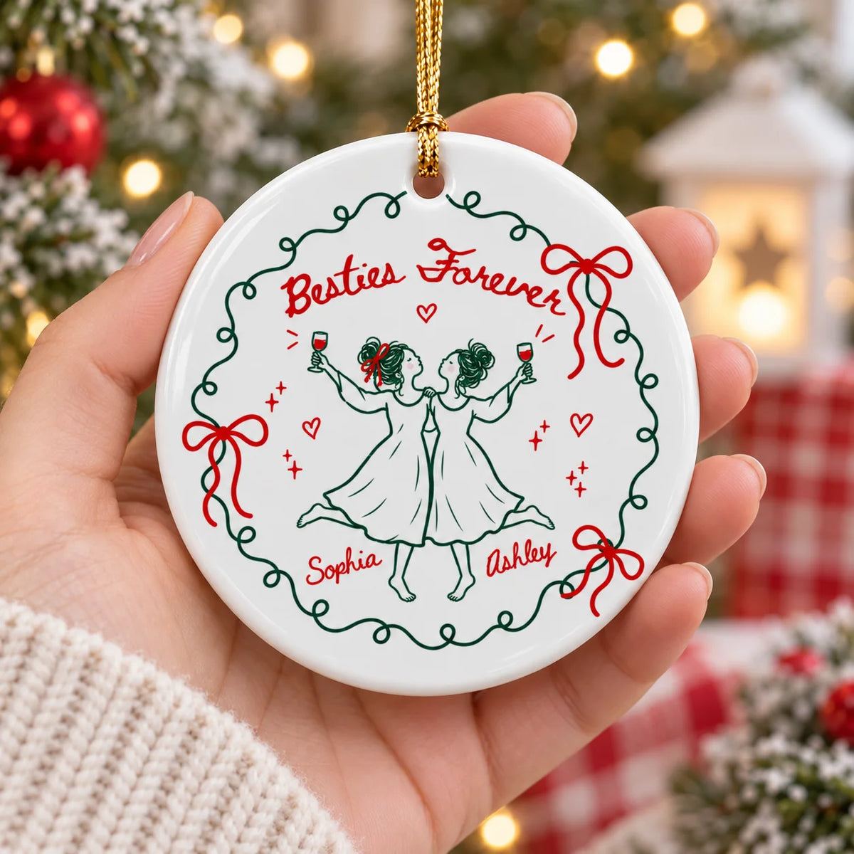 A person's hand holding a round ceramic Christmas ornament with custom names Sophia and Ashley in front of Christmas lights.
