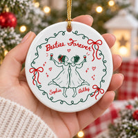 A person's hand holding a round ceramic Christmas ornament with custom names Sophia and Ashley in front of Christmas lights.