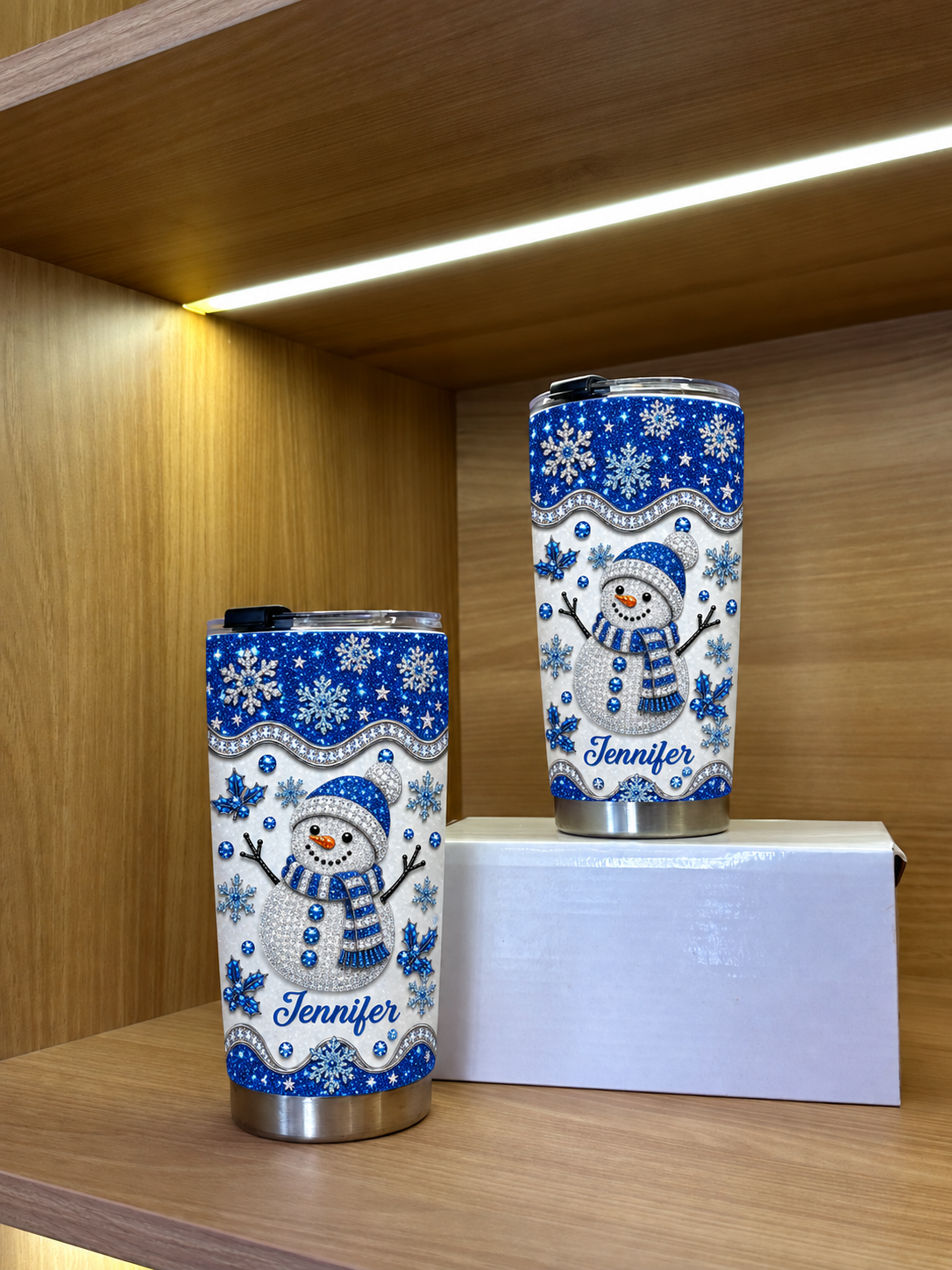 Two stainless steel winter tumblers with snowman design on a wooden shelf.