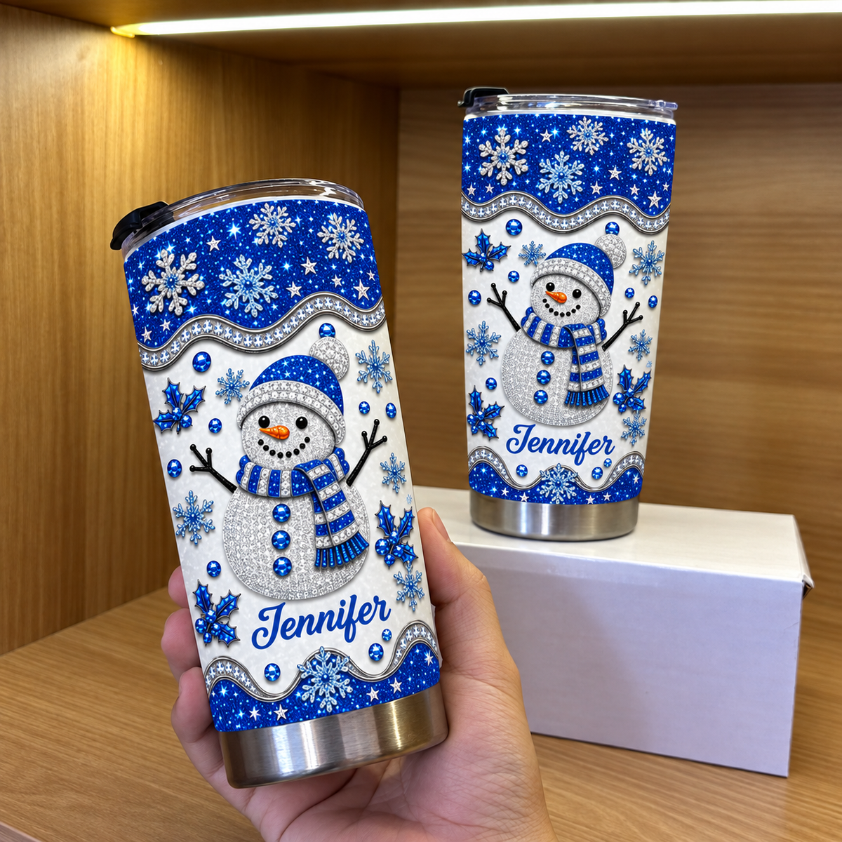 Hand holding blue glitter snowman tumbler next to another on box, wooden shelf.