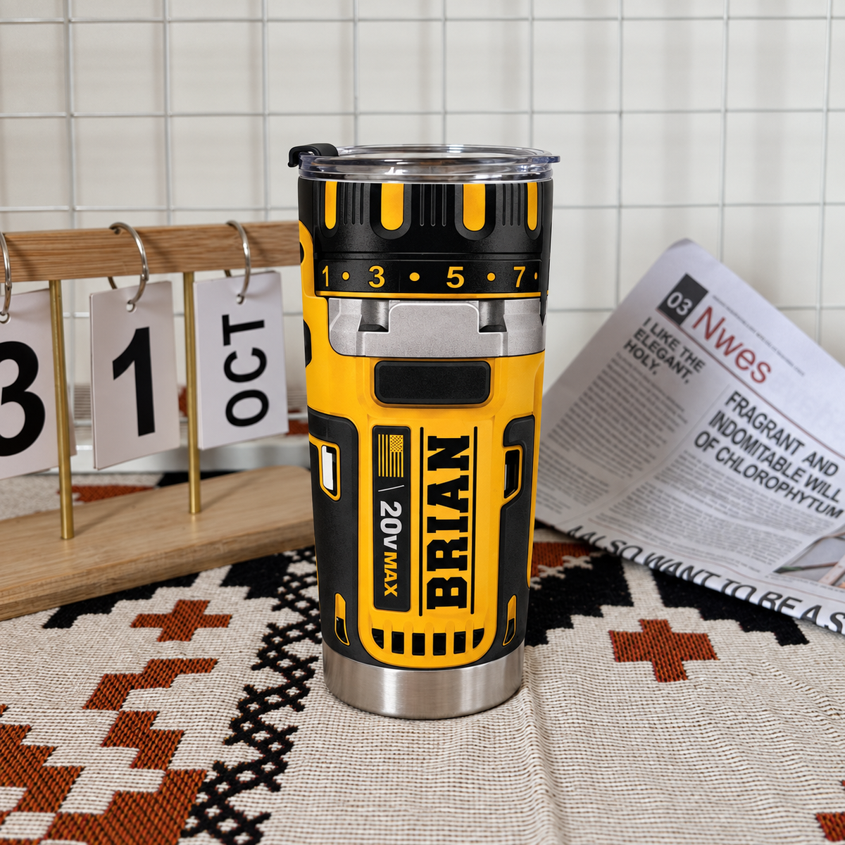Drill theme stainless steel insulated cup with name BRIAN placed on a patterned rug next to a desk calendar and newspaper.