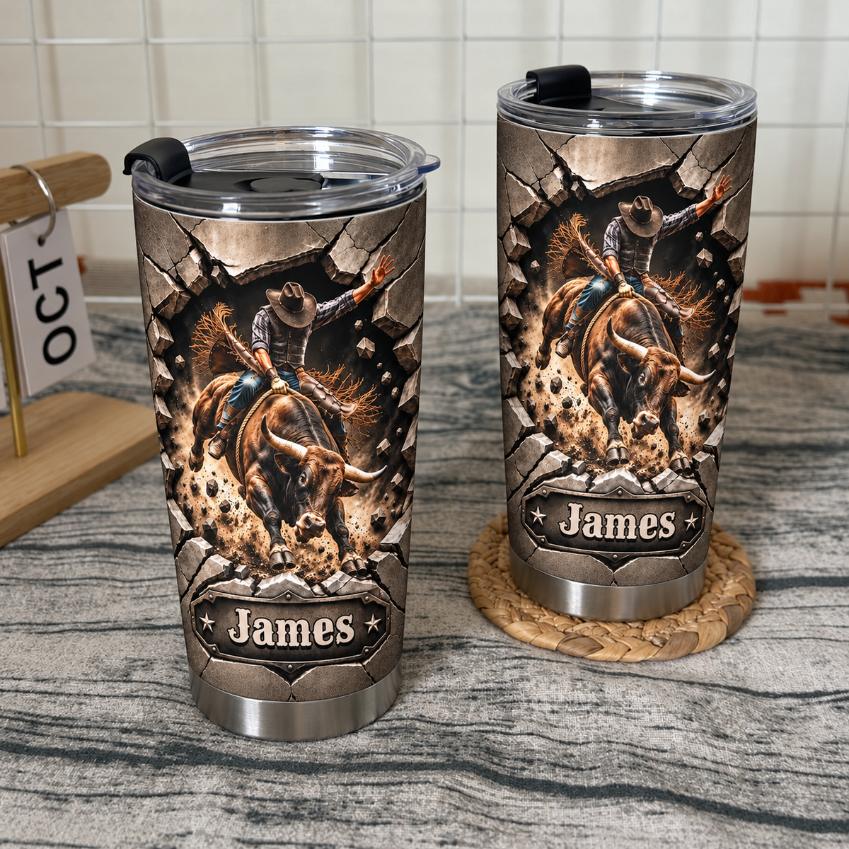 Two custom cowboy tumblers with a bucking bull design standing on a textured surface next to a wooden desk calendar.