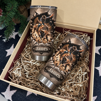 Two custom name rodeo tumblers nested inside an open gift box filled with paper shreds against a starry holiday backdrop.