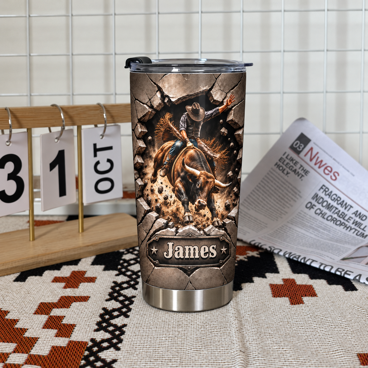 A personalized western rodeo gift tumbler standing on a patterned fabric mat next to a desktop calendar and newspaper.