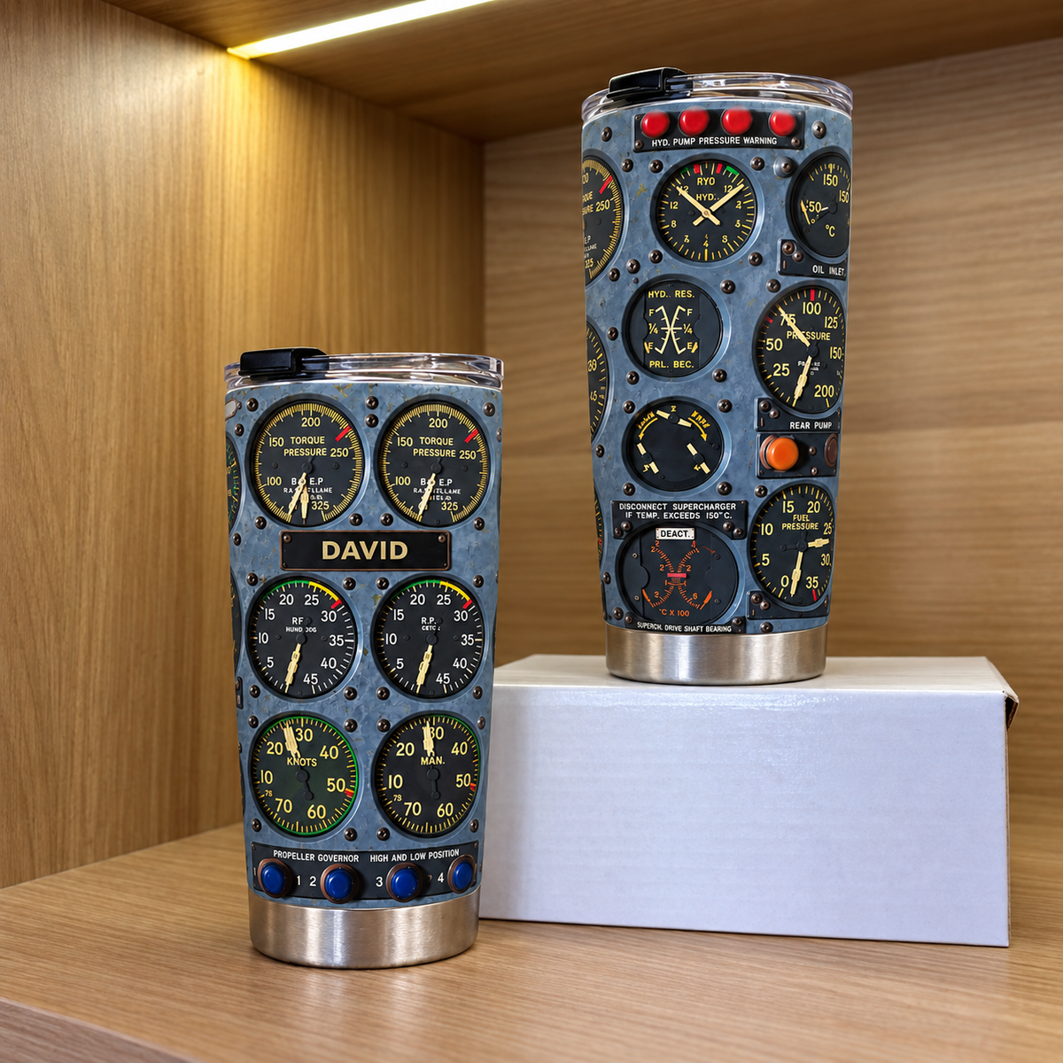 Two stainless steel cockpit instrument panel tumblers in different sizes displayed on a wooden shelf next to a white box.