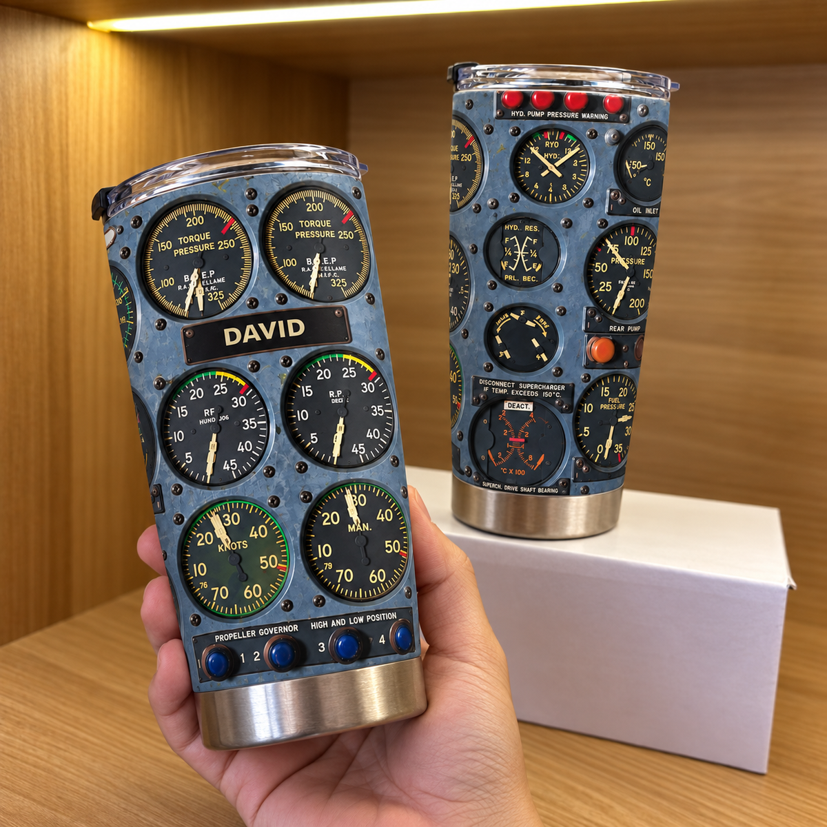 Hand holding a stainless steel cockpit instrument panel tumbler with custom name David against a wooden shelf.
