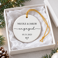 Personalized ceramic ornament resting inside an open white gift box lined with tissue paper next to pine needles.