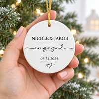 Hand holding a round white ceramic engagement ornament featuring custom names and date against a Christmas tree background.