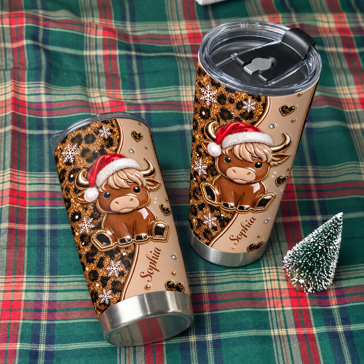 Two custom Christmas highland cow leopard print tumblers displayed on red and green plaid fabric with a mini tree.