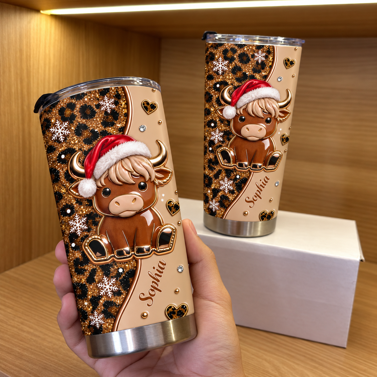 Hand holding Christmas highland cow leopard print tumbler with name Sophia, next to another on white box on wood shelf.