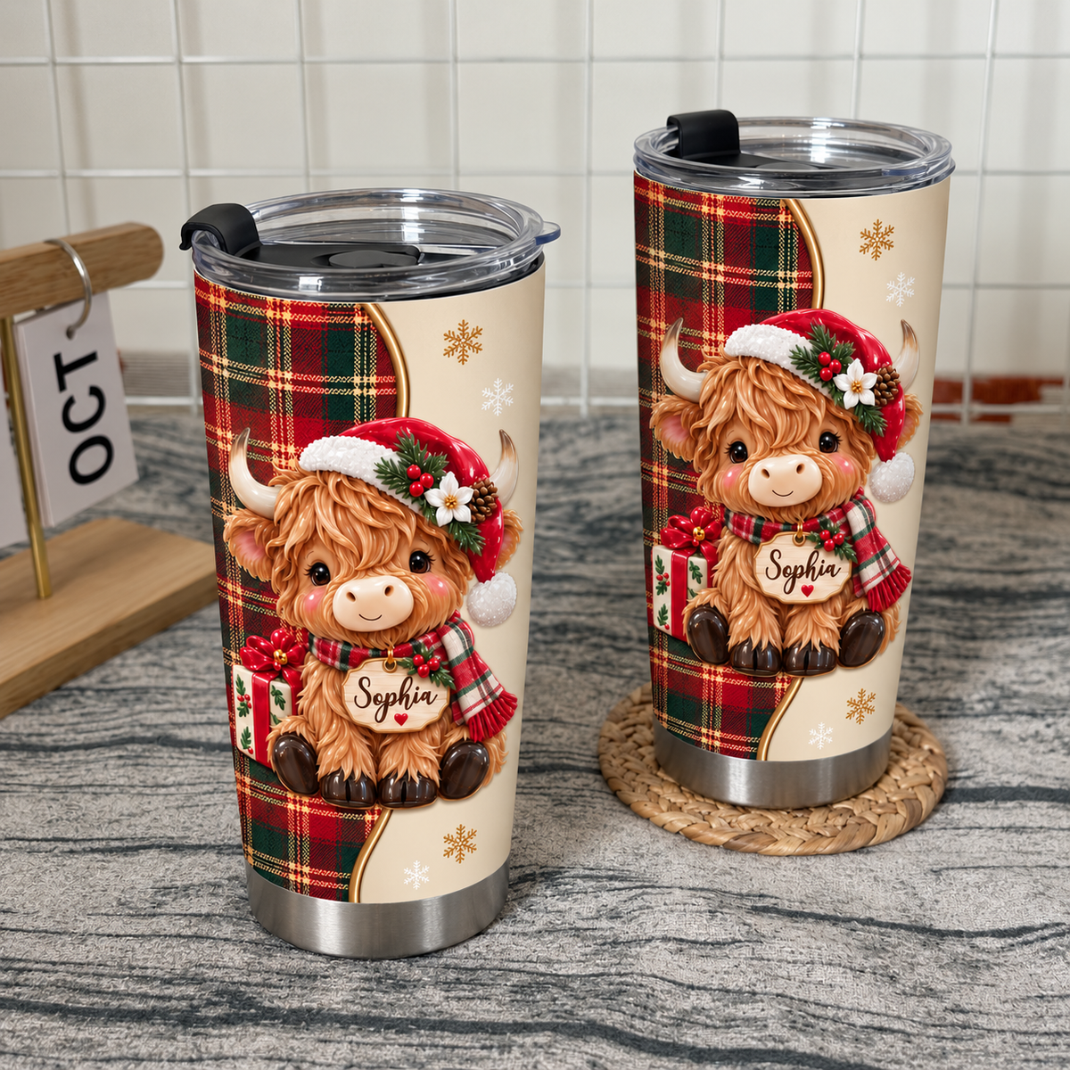 Two Christmas Highland cow tumblers standing on a grey textured surface next to a wooden block calendar showing October.