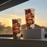 Two personalized Christmas Highland cow tumblers displayed on a wooden sill with a glowing sunset and silhouette view