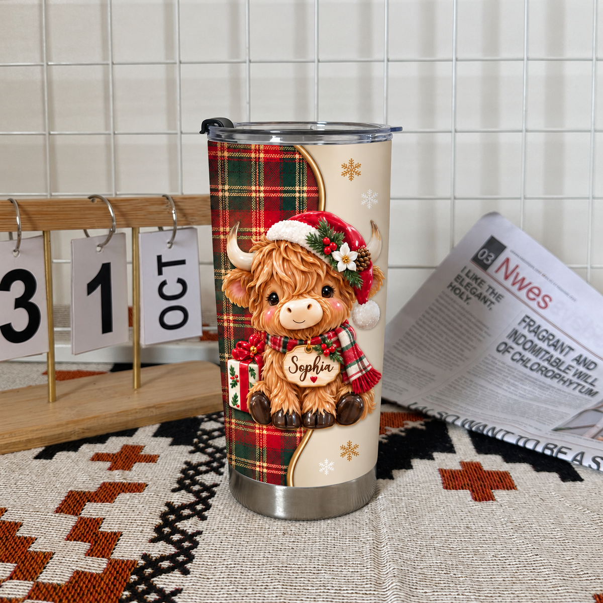 A Christmas Highland cow tumbler standing on a patterned rug next to a white wall grid, calendar, and open newspaper.