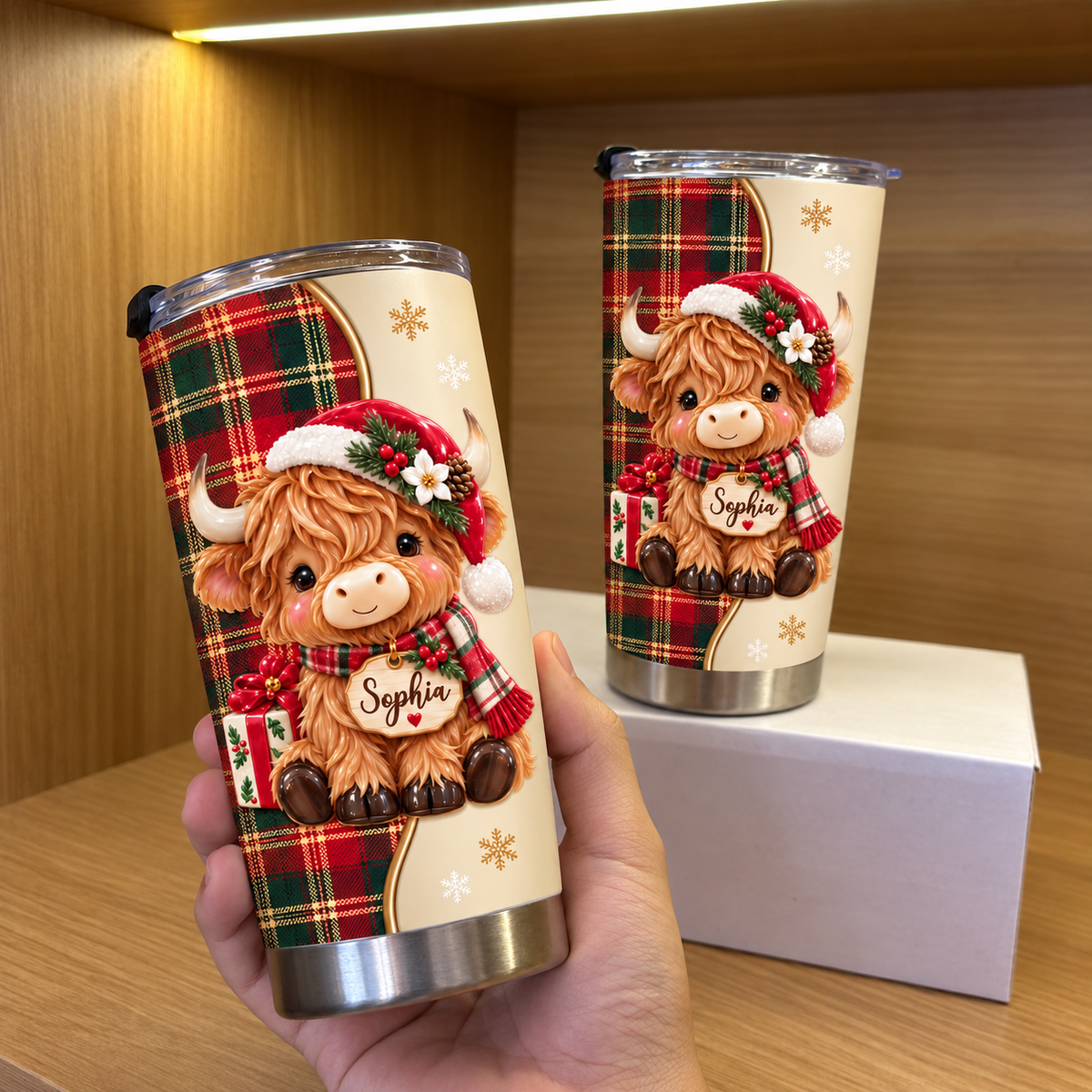 Two custom Christmas Highland cow tumblers held in a hand, showing red tartan plaid and a kawaii baby cow in a Santa hat.