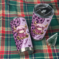 Two personalized purple cow tumblers laid across a green and red plaid fabric surface next to a miniature decorative