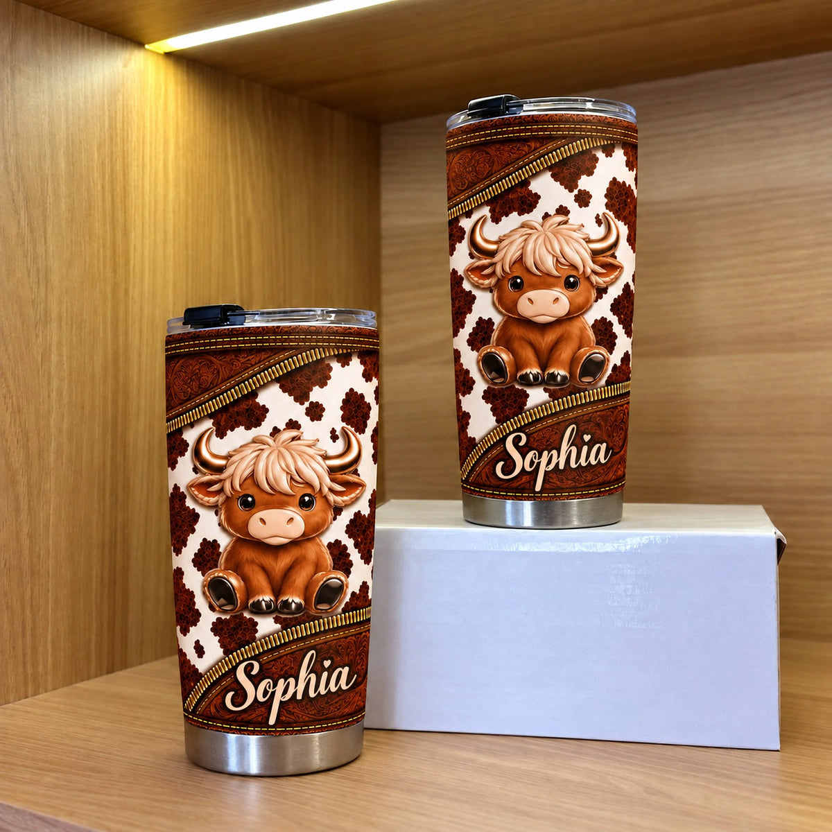 Two stainless steel travel mugs with cute Highland cow design displayed upright on a wooden shelf beside a white box