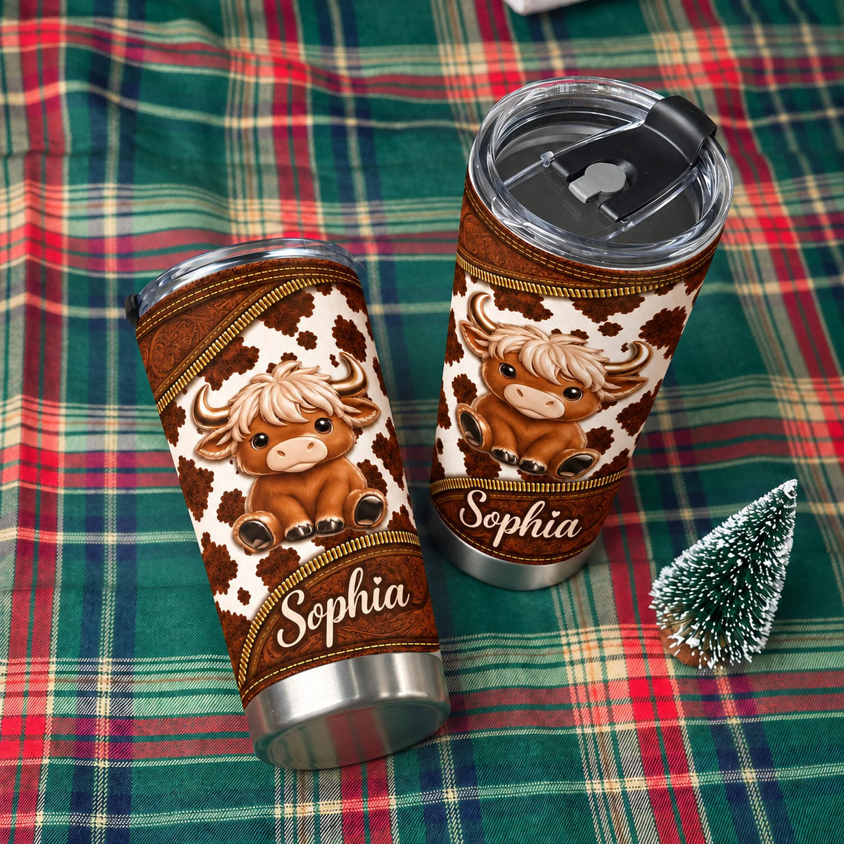 Two custom name tumblers with Highland cow print and leather-textured borders displayed on a green and red plaid cloth