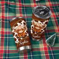 Two custom name tumblers with Highland cow print and leather-textured borders displayed on a green and red plaid cloth