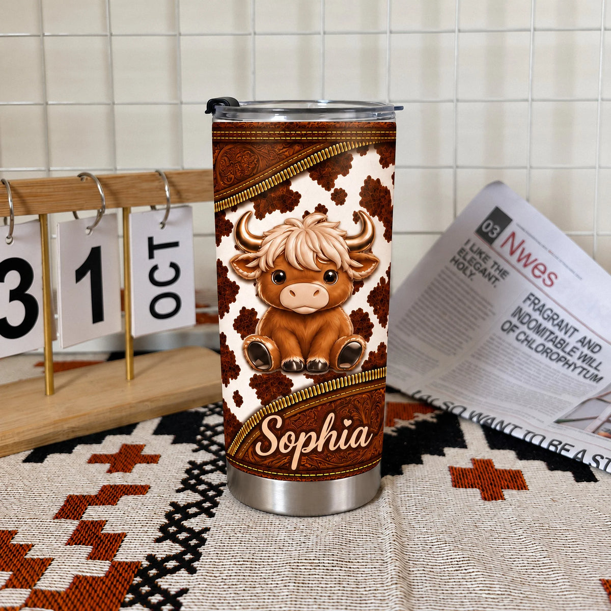 Stainless steel travel mug with cute Highland cow graphic placed on a rustic blanket next to a calendar and newspaper