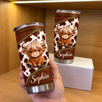 A hand holds up a personalized stainless steel travel mug with a cute Highland cow and custom name in front of a shelf