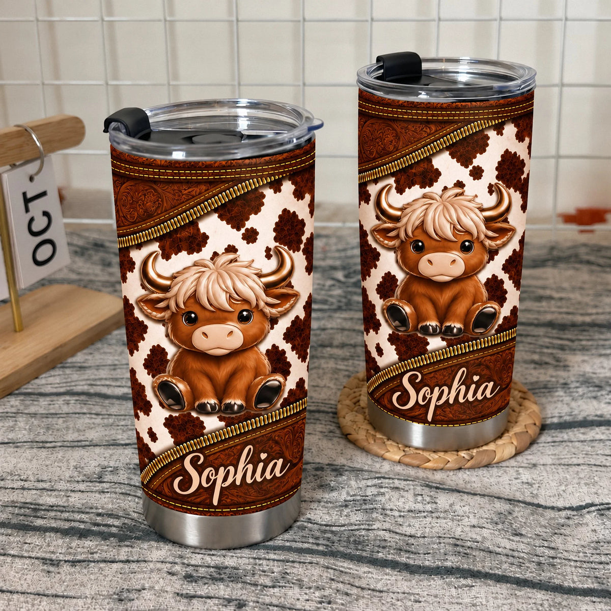 Two cute Highland cow tumblers with custom names displayed on a textured surface next to a small wooden calendar