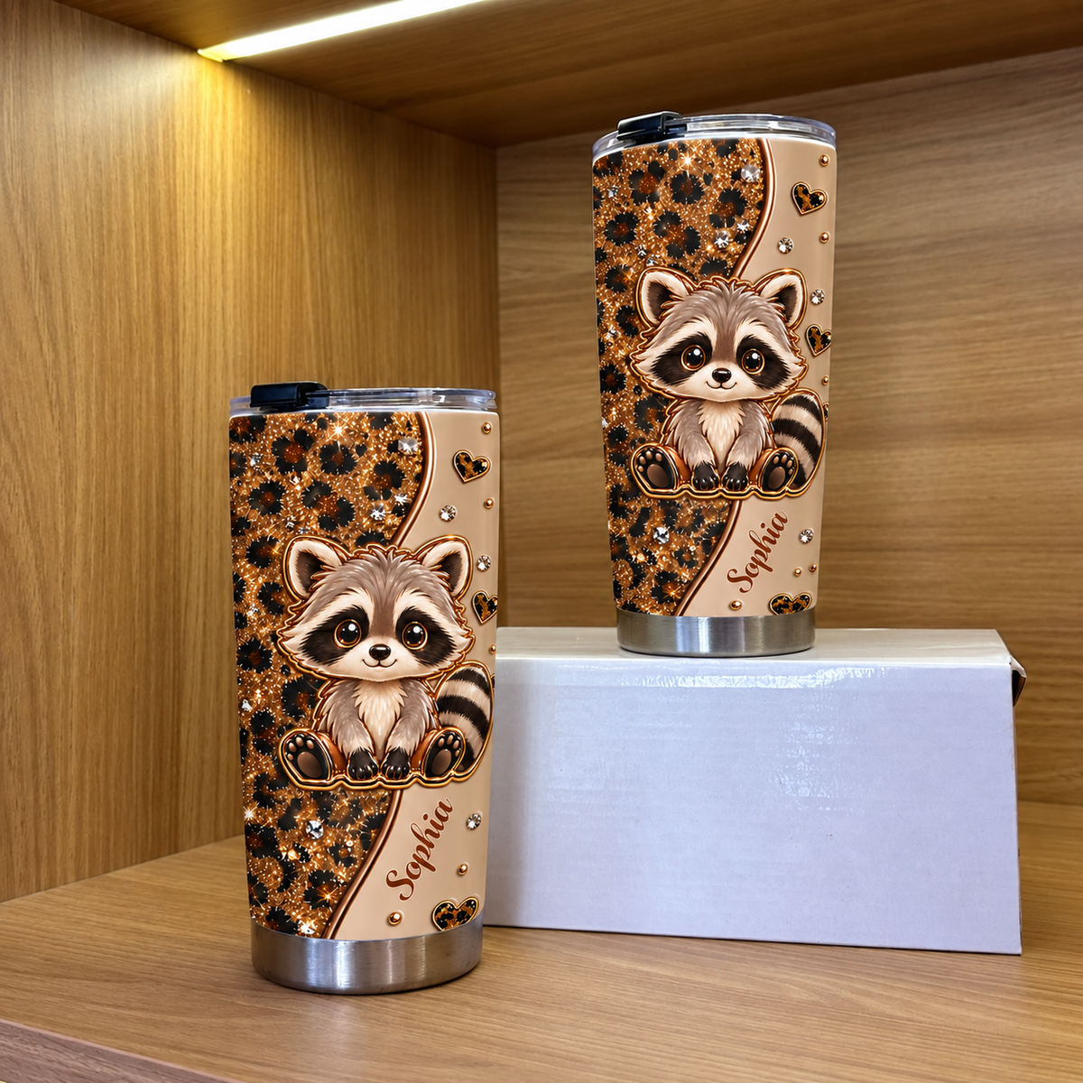 Two stainless steel tumblers with cute raccoon artwork and gold accents stand inside a wooden shelf with warm lighting.