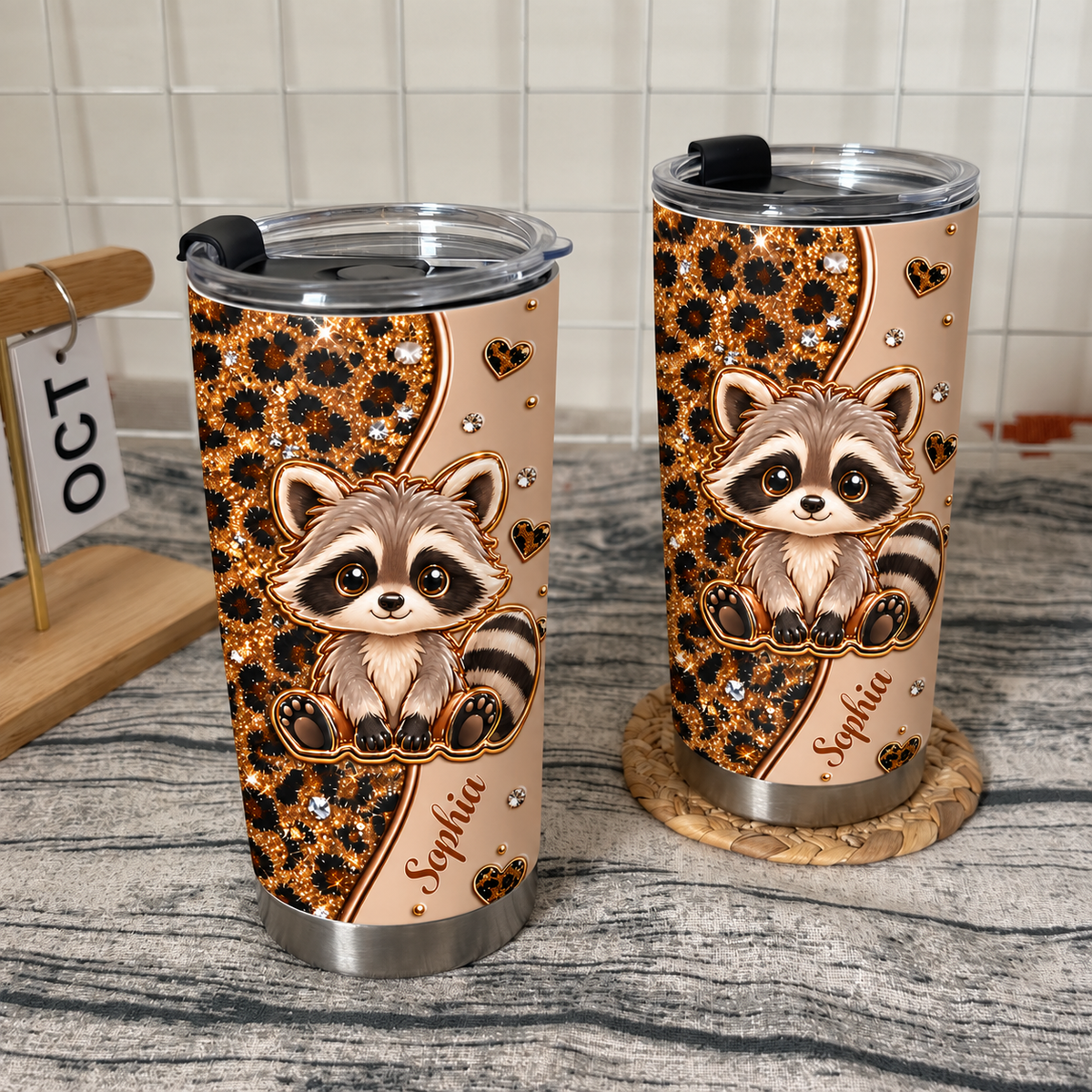 Two stainless steel drinkware tumblers featuring a raccoon and leopard print stand on a textured surface with a calendar.